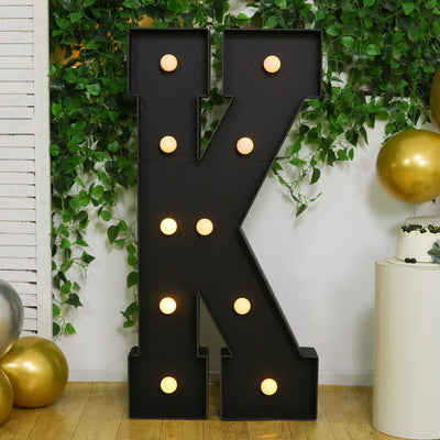 Black marquee letter "K" with lights against greenery and gold balloons.