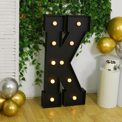 Black marquee letter 'K' with lights, surrounded by balloons and greenery.