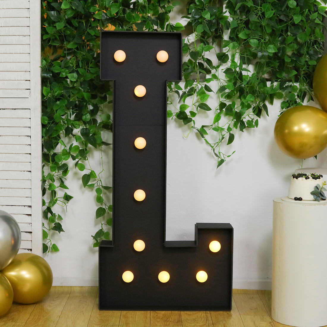 Large illuminated letter "L" decoration for events, surrounded by greenery and balloons.