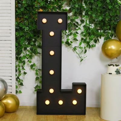 Large illuminated letter "L" decoration for events, surrounded by greenery and balloons.