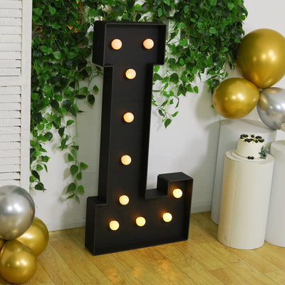 Large illuminated letter "L" marquee sign with lights, surrounded by greenery and balloons.