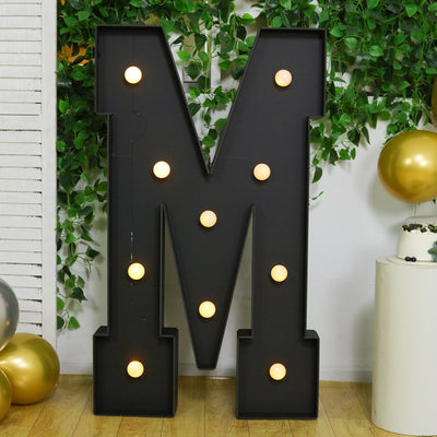 Large black marquee letter "M" with lights, styled with greenery and balloons.