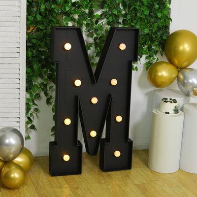 Large illuminated letter "M" marquee sign in a festive setting with balloons.