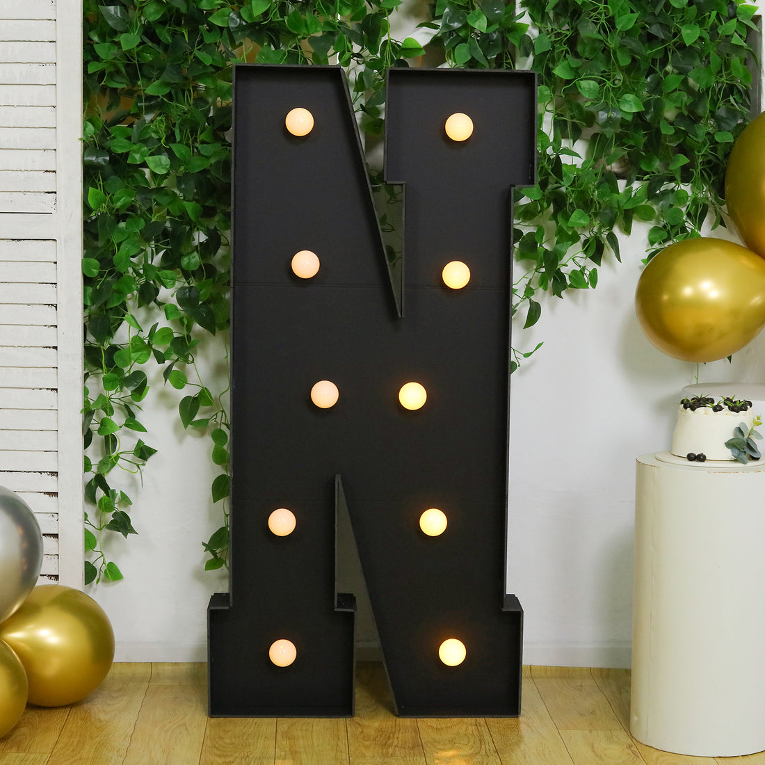 Light-up marquee letter "N" with bulbs, surrounded by greenery and balloons.
