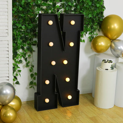 Black marquee letter "N" with lights, surrounded by green foliage and balloons.