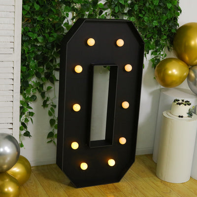 Large decorative marquee letter "O" with lights, set against greenery and balloons.