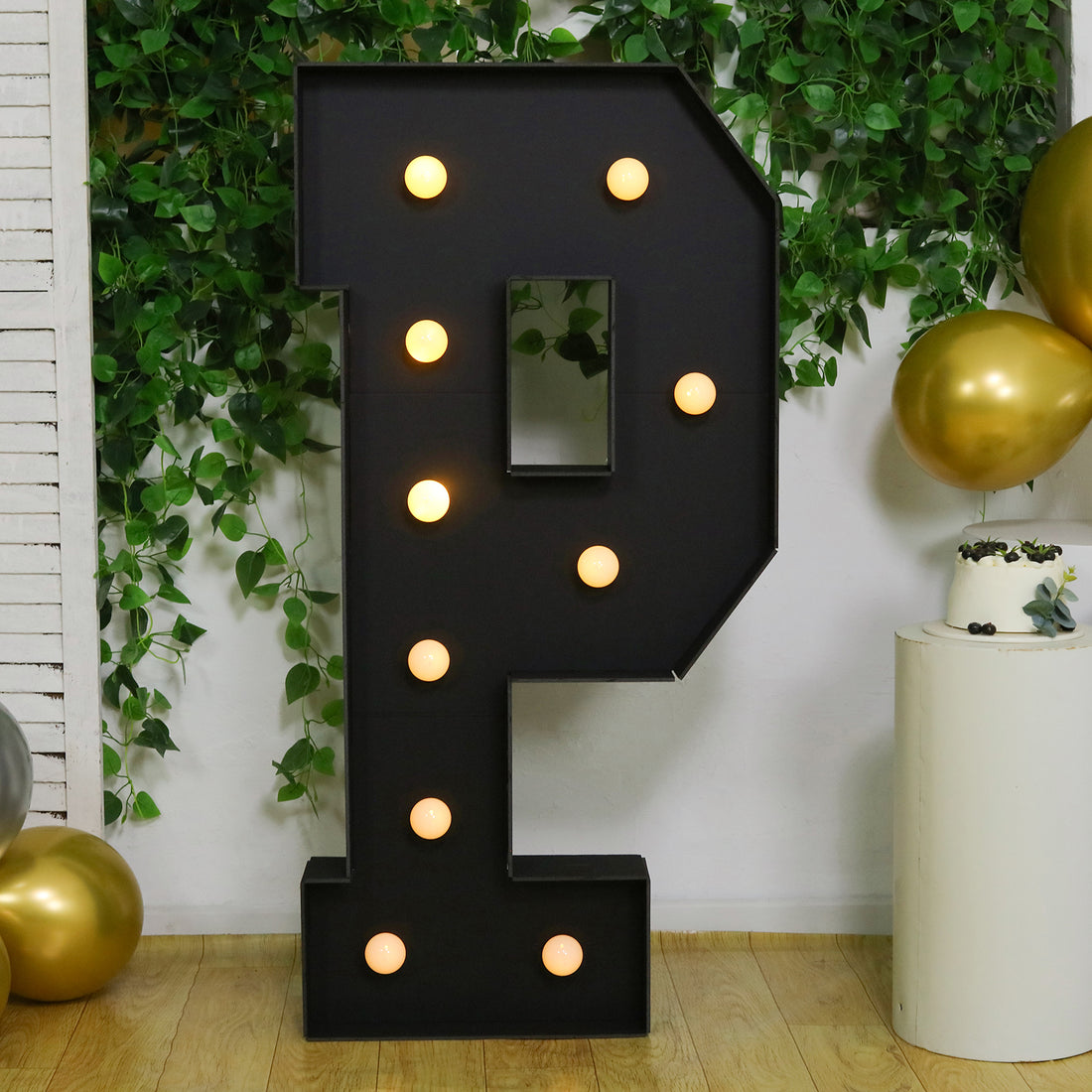 Large illuminated letter P with decorative lights, set against a greenery backdrop.