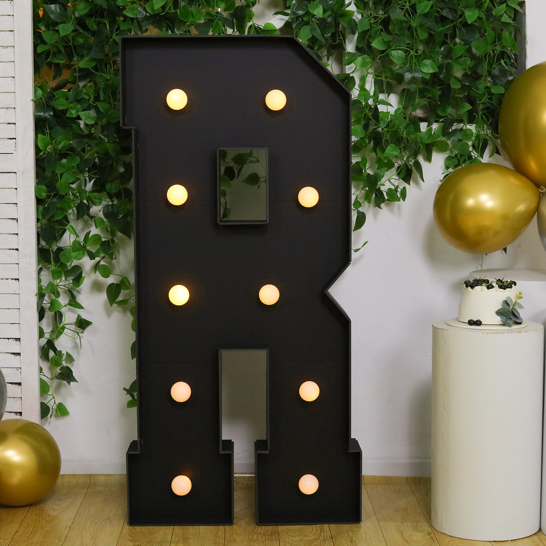 Black illuminated letter "R" with lights, surrounded by green foliage and gold balloons.