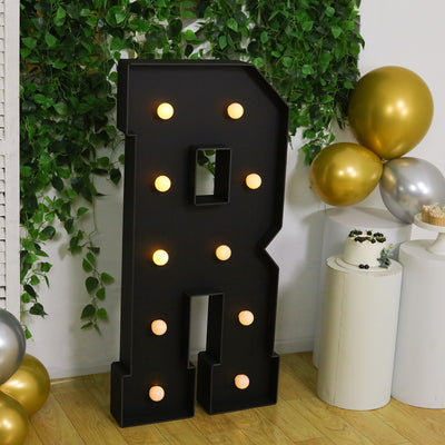 Black marquee letter "R" with lights, surrounded by greenery and balloons.