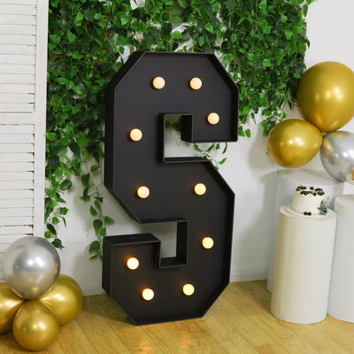 Large illuminated letter "S" with decorative lights, surrounded by greenery and balloons.