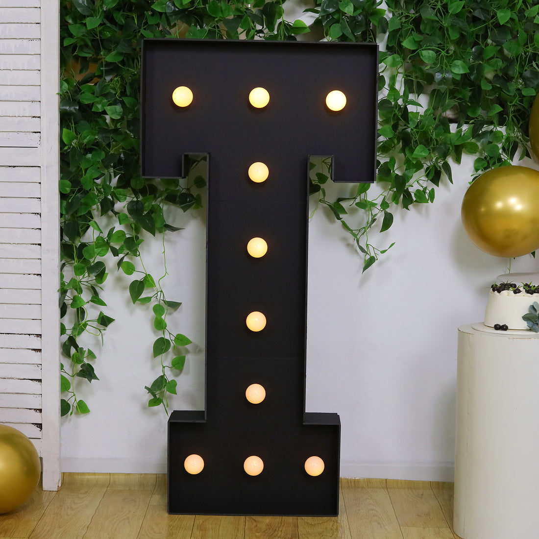 Black marquee letter T with lights, surrounded by greenery and gold balloons.