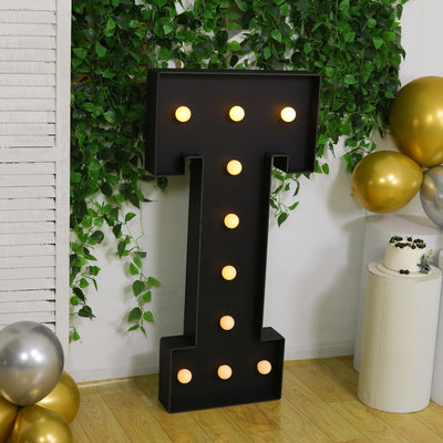 Black marquee letter "I" with lights, set against greenery and balloons.