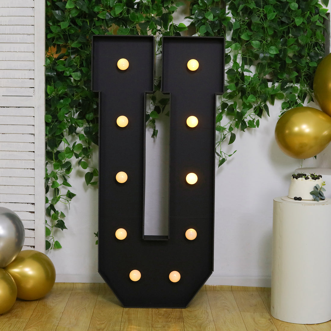Large illuminated letter "U" marquee sign with bulbs, set against greenery.