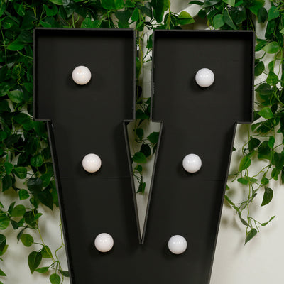 Black marquee letter 'V' with white bulbs on green foliage backdrop.