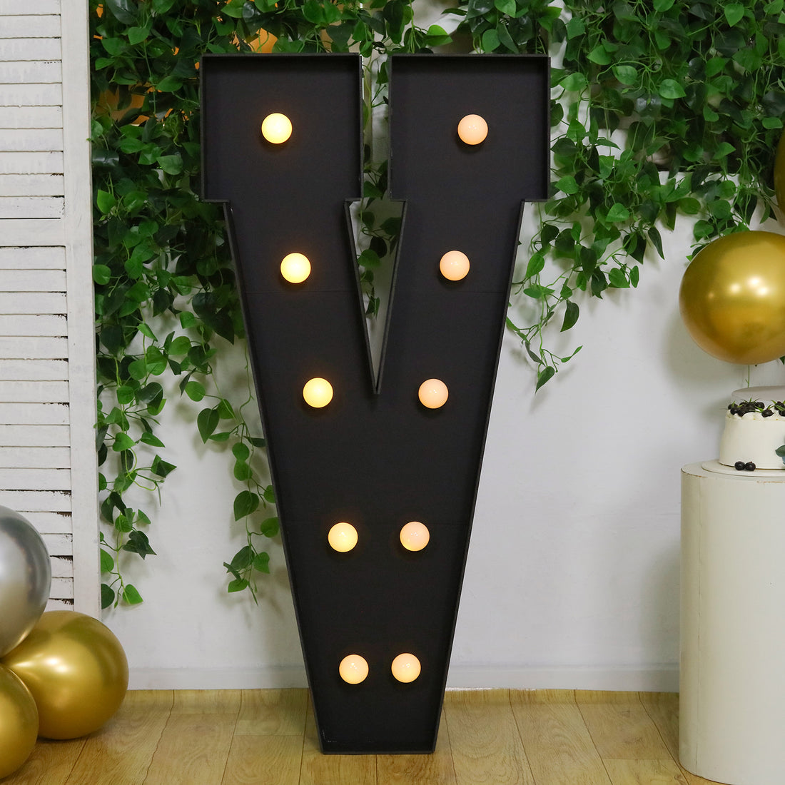 Large decorative letter V with lights, set against a green plant backdrop.