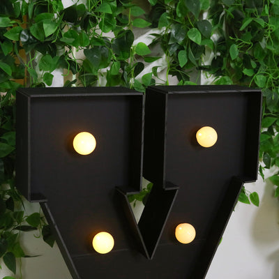 Vintage marquee letter "V" with warm glow bulbs against greenery backdrop.
