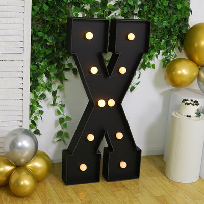 Large decorative black letter "X" with lights, surrounded by balloons and greenery.