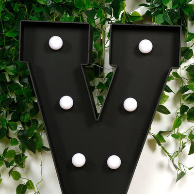 Black marquee letter "V" with white bulbs surrounded by green foliage.