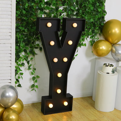 Large illuminated letter "Y" in black with bulbs, surrounded by greenery and balloons.
