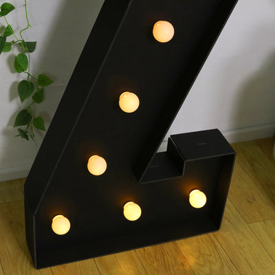 Black marquee letter with warm white bulbs illuminated on a wooden floor.