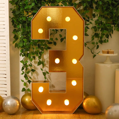 Gold marquee number three with lights and greenery backdrop.