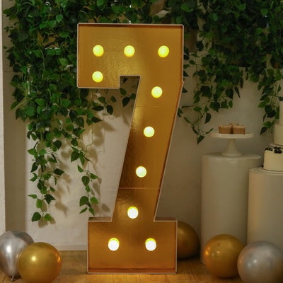 Gold marquee number 7 with lights, surrounded by balloons and greenery.