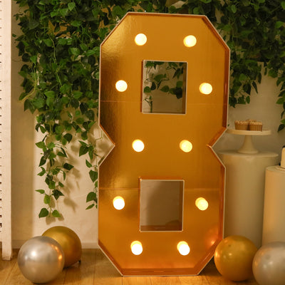 Gold marquee number 8 with lights against greenery backdrop.