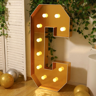 Gold marquee letter C with lights, surrounded by greenery and party decor.