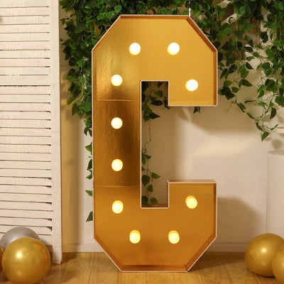 Gold marquee letter "C" with lights against greenery backdrop.