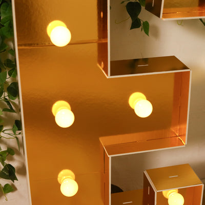Gold marquee letter with warm glowing bulbs and green foliage accents.
