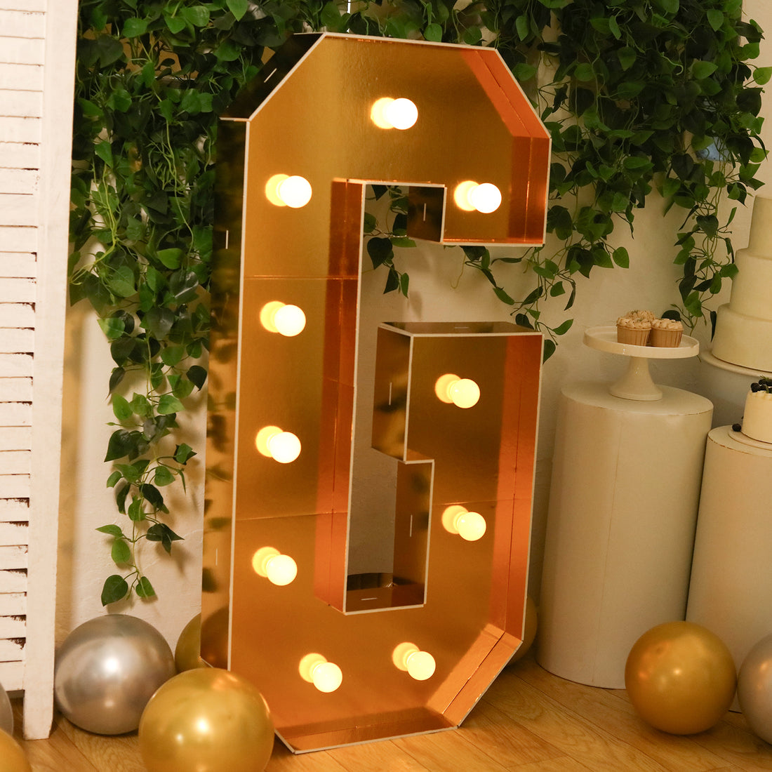 Gold marquee letter G with lights, surrounded by greenery and balloons.