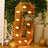 Gold marquee letter G with lights, surrounded by greenery and balloons.