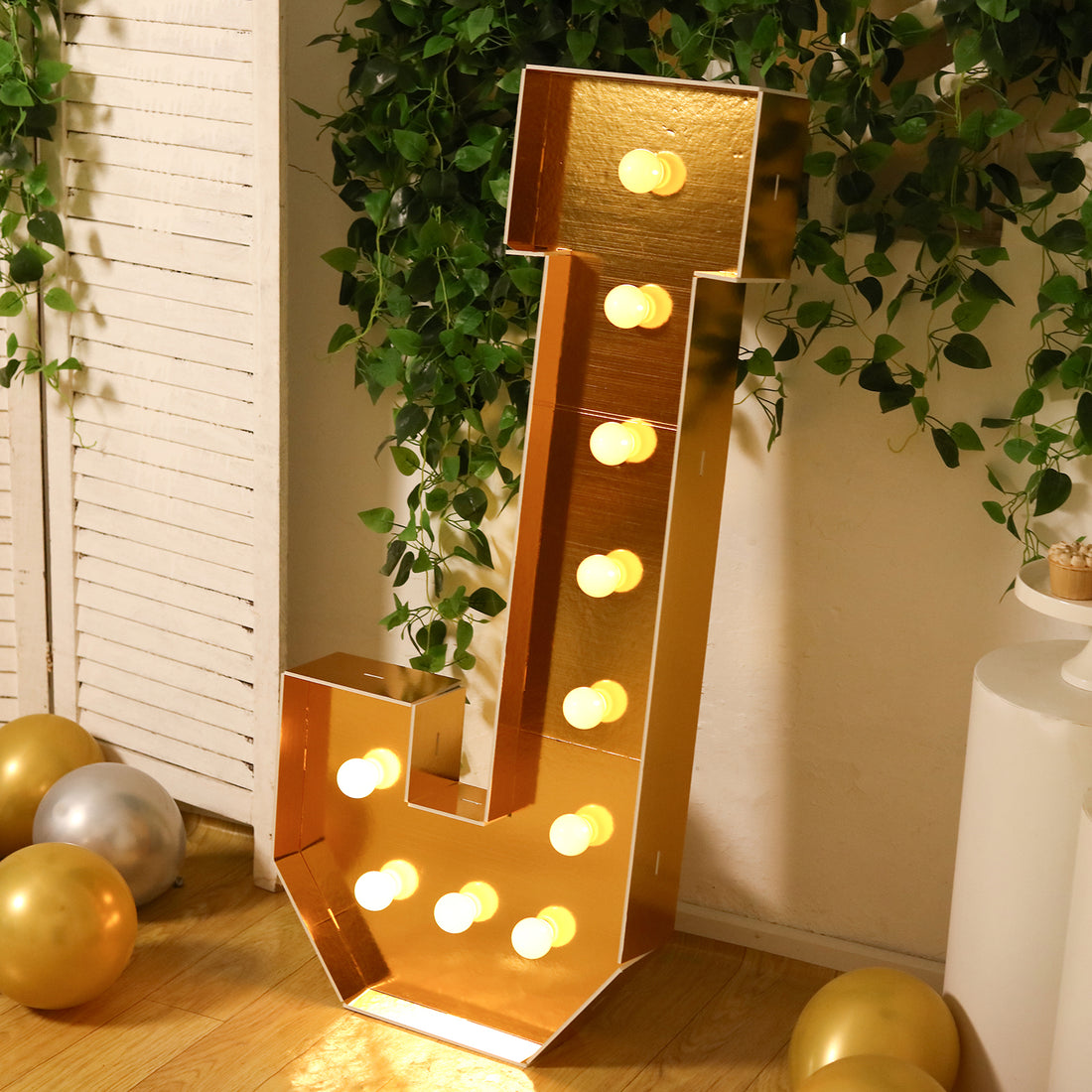 Gold marquee letter "J" illuminated with bulbs, surrounded by greenery and balloons.