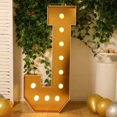 Gold marquee letter "J" with lights, set against greenery and decorative elements.