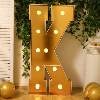 Gold marquee letter "K" with lights, set against greenery and decor.