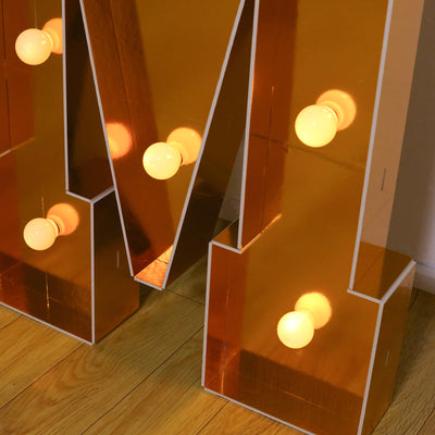 Golden decorative letter "M" with light bulbs on wooden floor.
