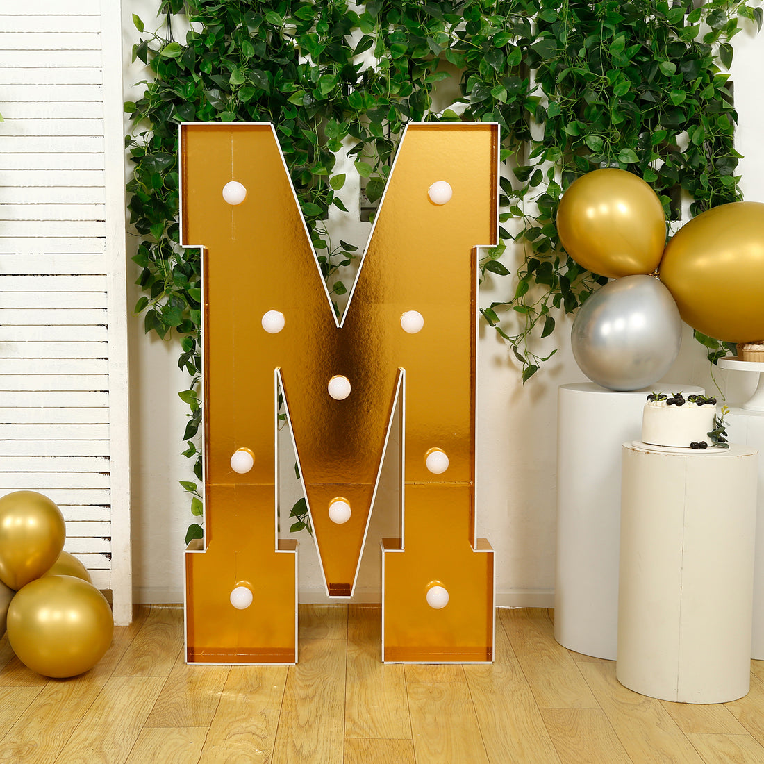 Gold marquee letter "M" with lights, surrounded by greenery and balloons.