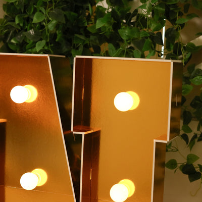 Golden marquee letter with bulbs, reflecting light against greenery.