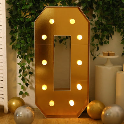 Gold marquee number zero with lights, surrounded by balloons and greenery.