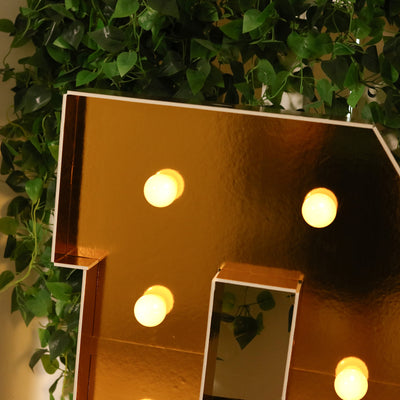 Gold marquee letter with light bulbs, surrounded by greenery.