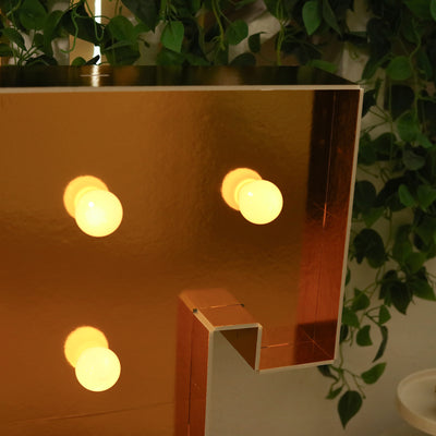 Gold letter light with four glowing bulbs against a background of greenery.