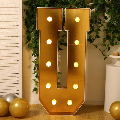 Gold marquee letter "U" with lights, surrounded by greenery and balloons.