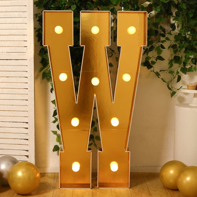 Gold marquee letter "W" with lights, set against greenery and balloons.