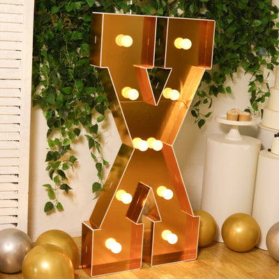 Gold marquee letter X with lights, surrounded by greenery and balloons.