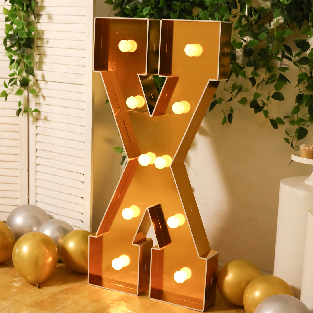 Gold marquee letter "X" with lights, decorated with balloons and greenery.