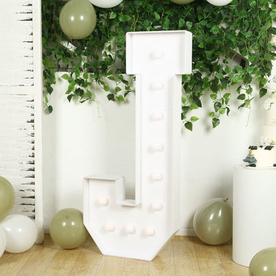 White decorative letter "J" with lights, surrounded by greenery and balloons.