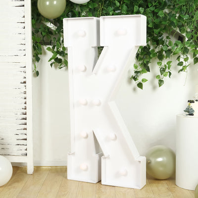 Large decorative letter "K" with lights, set against greenery backdrop.