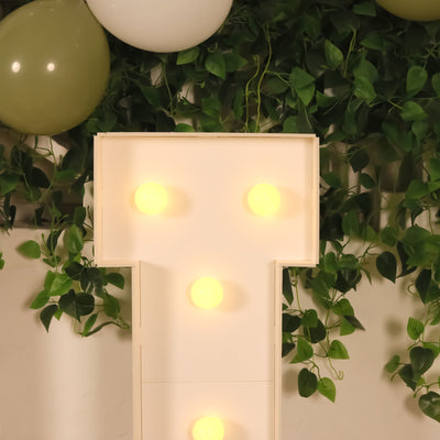 Illuminated letter T marquee sign with warm bulbs and greenery backdrop.