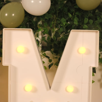 Light-up decorative letter "M" with bulbs, surrounded by greenery and balloons.