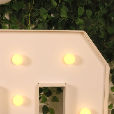Decorative light-up letter with bulbs against greenery backdrop.